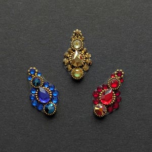 May include: Three decorative jewel embellishments in gold, blue, and red. The gold piece has amber and green stones. The blue piece features sapphire and teal stones. The red piece has ruby and orange stones, all on a black background.