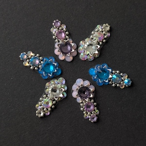 May include: Six bejeweled hair clips arranged in a circular pattern. The clips feature blue, purple, and clear gemstones set in silver-toned metal. The gemstones are arranged in floral and cluster designs. The clips are displayed on a dark background.
