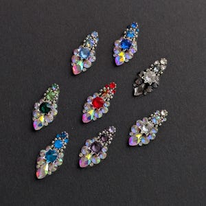 May include: Eight decorative rhinestone embellishments in various colours, including blue, green, red, and purple. Each piece features a central gemstone surrounded by smaller, iridescent rhinestones, arranged on a dark background.