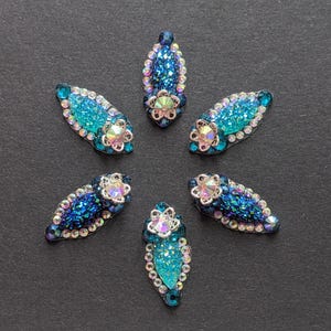 May include: Seven jewel-toned decorative embellishments arranged on a dark grey surface. Each piece features a central flower-shaped rhinestone and is surrounded by iridescent, sparkling crystals in shades of blue and teal. Ideal for craft projects.