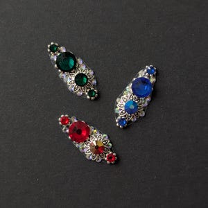 May include: Three decorative hair clips with jewel-toned gemstones. Each clip features a central large gem surrounded by smaller stones and a floral design. The clips are in shades of green, blue, and red, with silver-coloured metalwork.