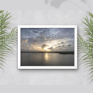May include: A framed photograph of a sunset over a body of water. The sky is a mix of blue, gray, and white clouds with the sun shining through. The water is calm and reflective, with a slight ripple.