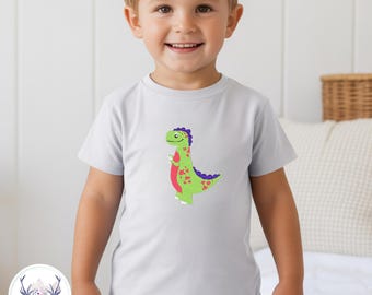May include: A light gray short-sleeved t-shirt featuring a centered graphic of a green dinosaur with a red belly, purple spikes, and red spots. The dinosaur has a smiling expression and is positioned on the front of the shirt.