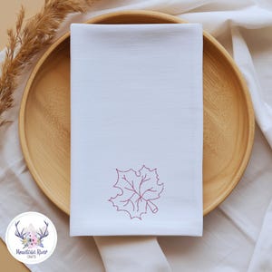 May include: A rectangular white cloth napkin featuring a single embroidered dark red maple leaf design centered near the bottom edge. The fabric has a smooth, woven texture with clean, straight stitched hems along all four sides of the textile.