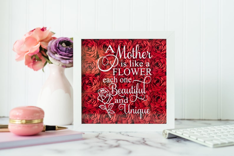 May include: A white framed artwork featuring a red rose design and the quote, "A Mother is like a Flower, each one Beautiful and Unique." A pink vase with flowers and a pink macaron box are in the foreground.
