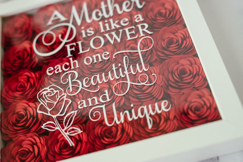 May include: A framed artwork with a background of red paper roses. White script reads, "A Mother is like a FLOWER, each one BEAUTIFUL and UNIQUE." A white rose outline is also present.