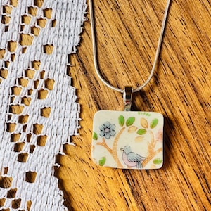 May include: A rectangular pendant necklace featuring a cream-colored ceramic base with a painted illustration of a small bird perched on a tree branch with green leaves and a blue flower, suspended from a thin silver-toned metal chain.