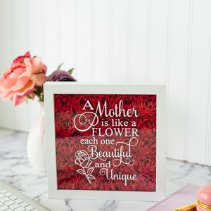 May include: A white-framed artwork featuring the text "A Mother is like a FLOWER each one Beautiful and Unique" over a bed of red roses. The frame rests on a marble surface, with a keyboard and flowers visible.