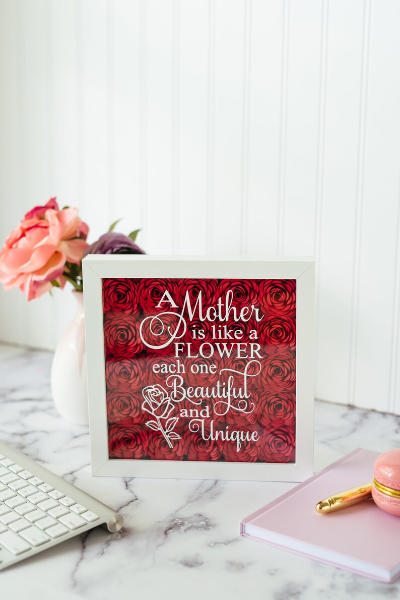May include: A white-framed artwork featuring the text "A Mother is like a FLOWER each one Beautiful and Unique" over a bed of red roses. The frame rests on a marble surface, with a keyboard and flowers visible.