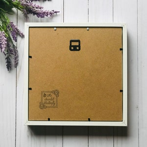 May include: The reverse of a white framed artwork, featuring a brown backing and a black metal hanger. The bottom left corner displays a stamp reading "The Scarlett Butterfly". Purple flowers are arranged in the upper left corner of the image.