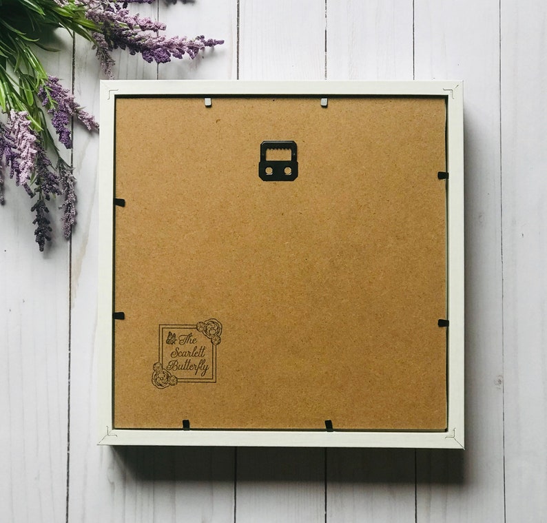May include: The reverse of a white framed artwork, featuring a brown backing and a black metal hanger. The bottom left corner displays a stamp reading "The Scarlett Butterfly". Purple flowers are arranged in the upper left corner of the image.