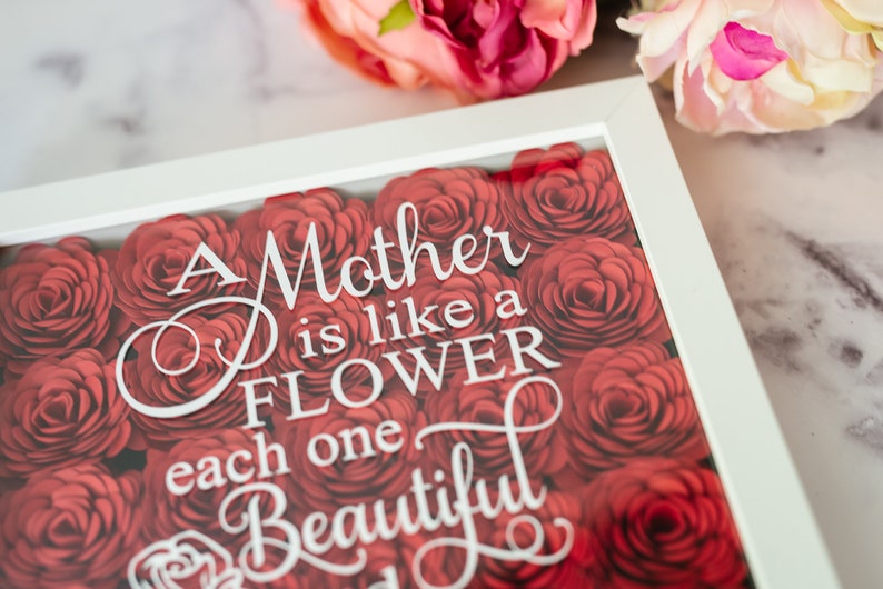 May include: A white-framed artwork with a floral design. The image features a bed of red roses, with the words "A Mother is like a FLOWER, each one Beautiful" in white script. Pink flowers are visible in the background.