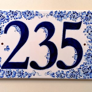 May include: A white ceramic house number sign with a blue floral border and the number 235 painted in blue.