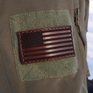 Op de afbeelding: Een close-up van een legerjas in olijfgroen, met een rechthoekige leren patch met het Amerikaanse vlagontwerp. De vlag is in bruin- en roodtinten, met een donkere achtergrond. De jas heeft een klittenband patch en een rits.