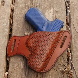 Könnte beinhalten: Eine blaue Handfeuerwaffe liegt auf einem braunen Lederholster mit einem gewebten Design. Das Holster mit zwei Schlitzen befindet sich auf einer verwitterten Holzoberfläche.