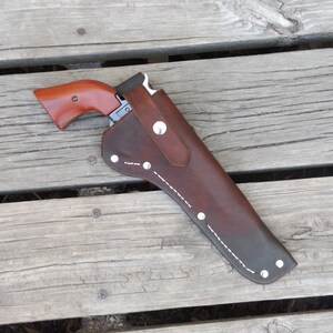 May include: A brown leather holster with a silver button and white stitching, holding a handgun with a wooden handle. The holster is on a wooden surface.