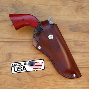 May include: A revolver with a red wooden grip is partially holstered in a brown leather holster. The holster is secured with metal rivets and a snap closure. A "MADE IN USA" sticker is visible.