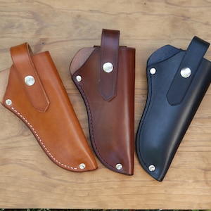May include: Three leather knife sheaths in shades of tan, brown, and black. Each sheath has a loop for a belt and is secured with a silver snap. The sheaths are stitched with white thread and are displayed on a wooden surface.