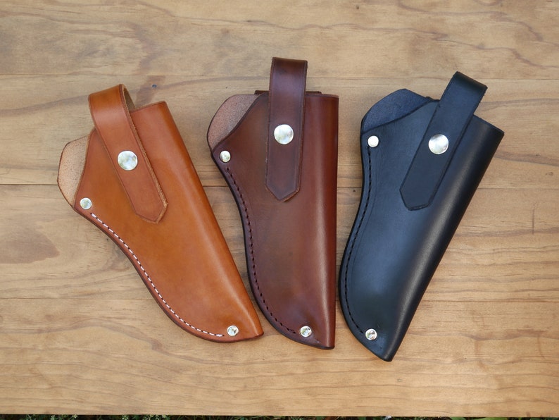 May include: Three leather knife sheaths in shades of tan, brown, and black. Each sheath has a loop for a belt and is secured with a silver snap. The sheaths are stitched with white thread and are displayed on a wooden surface.