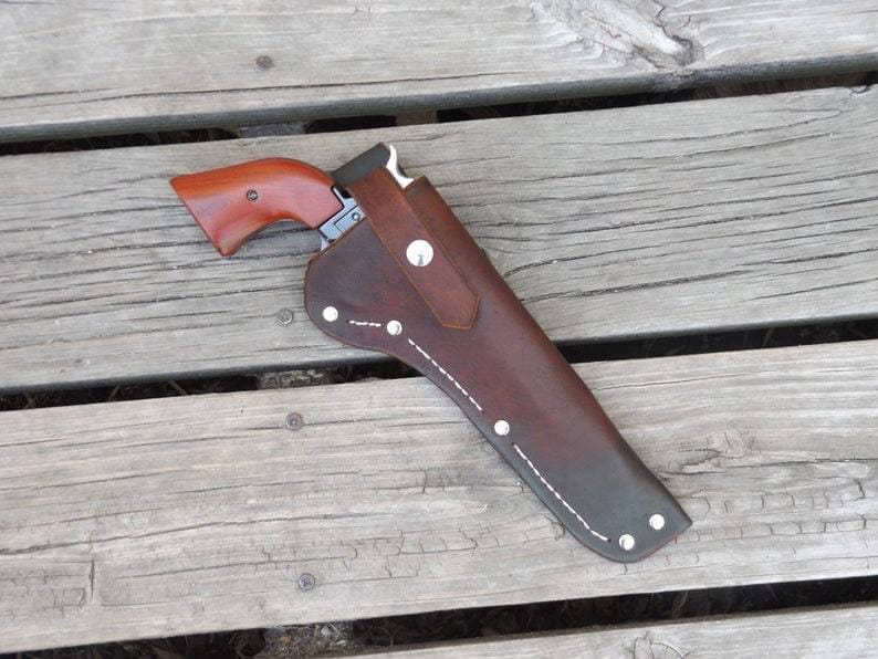 May include: A brown leather holster with a silver-colored revolver. The revolver has a wooden handle. The holster is stitched with white thread and secured with silver rivets. The gun and holster are resting on a wooden surface.
