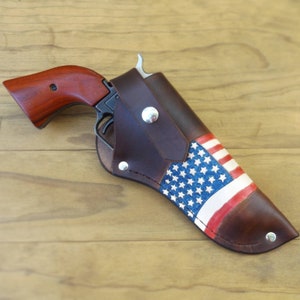 May include: A brown leather holster with an American flag design, holding a revolver with a wooden handle. The holster features a painted flag with blue stars and red and white stripes. Silver rivets secure the holster.
