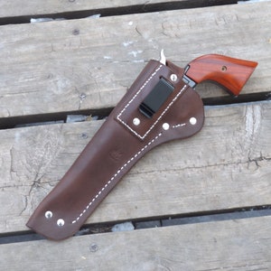 May include: A brown leather holster with white stitching and silver rivets, holding a handgun with a wooden grip. The holster has a black clip and is set against a weathered wooden surface.