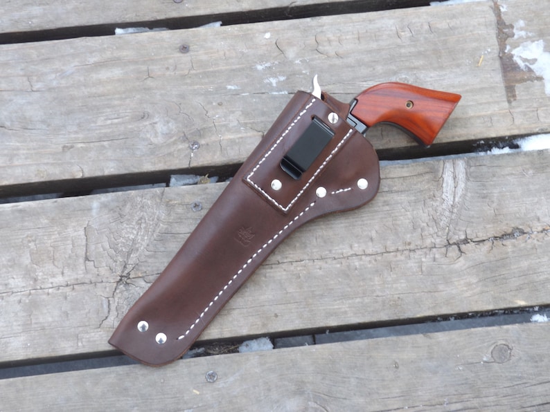 May include: A brown leather holster with white stitching and silver rivets, holding a handgun with a wooden grip. The holster has a black clip and is set against a weathered wooden surface.