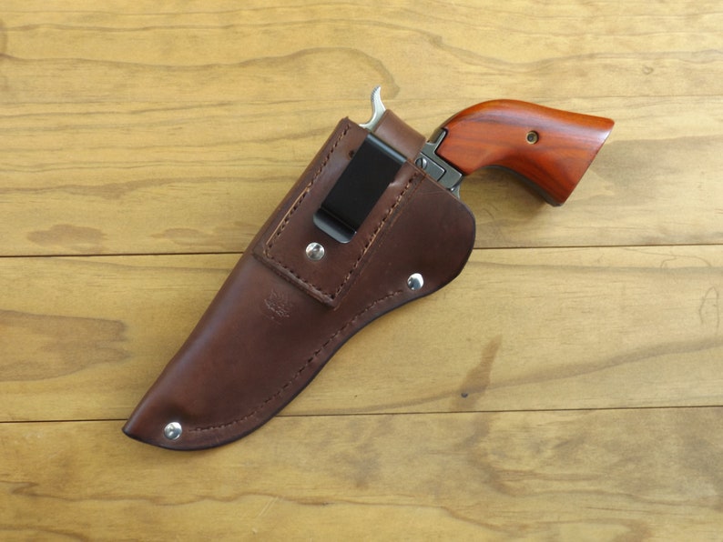 May include: A brown leather holster with a black metal clip holds a small handgun with a wooden grip. The gun is partially visible, with a silver trigger and barrel. The holster is secured with metal rivets.