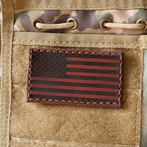 May include: A rectangular leather patch featuring the American flag design. The flag has a dark background with red stripes and white stars. The patch is stitched onto a camouflage fabric.
