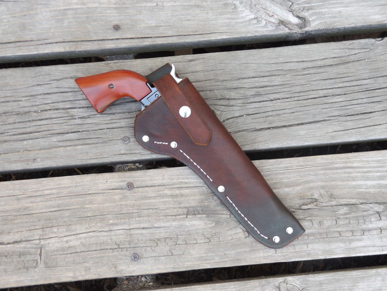 May include: A brown leather holster with a silver button and white stitching, holding a handgun with a wooden handle. The holster is on a wooden surface.