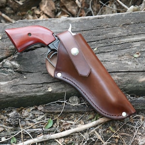May include: A brown leather holster with a silver button closure, holding a revolver with a dark metal frame and a reddish-brown wooden handle. The holster is stitched with a contrasting thread.