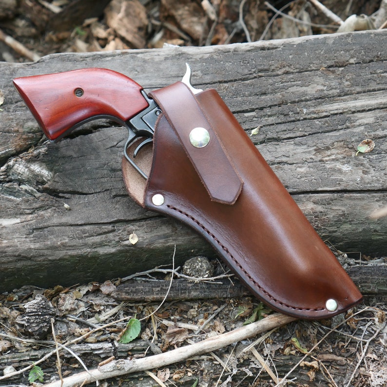 May include: A brown leather holster with a silver button closure, holding a revolver with a dark metal frame and a reddish-brown wooden handle. The holster is stitched with a contrasting thread.