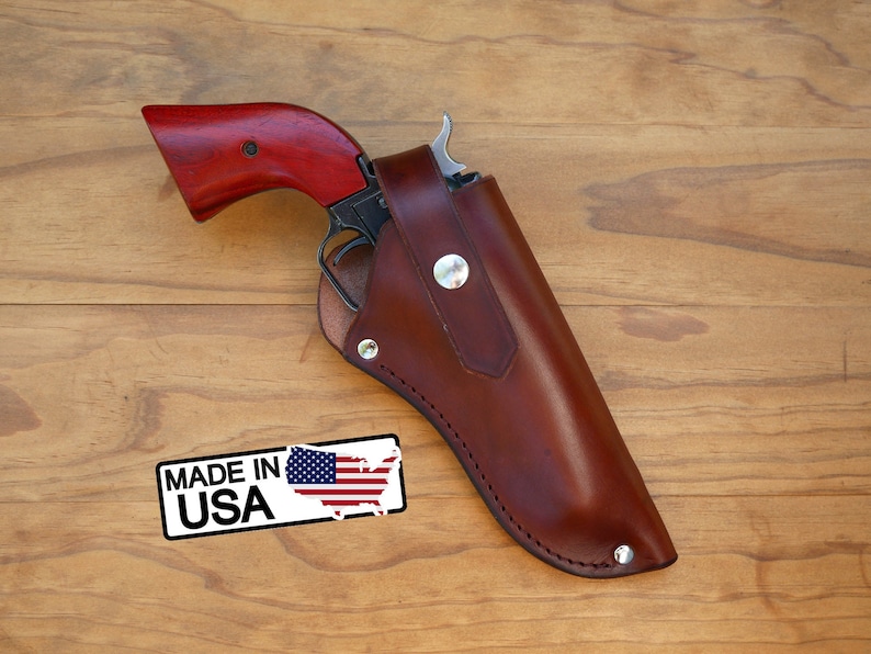 May include: A revolver with a red wooden grip is partially holstered in a brown leather holster. The holster is secured with metal rivets and a snap closure. A "MADE IN USA" sticker is visible.