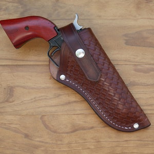 May include: A brown leather holster with a woven pattern holds a revolver with a dark metal frame and a reddish-brown wooden grip. The holster is secured with silver-colored rivets and white stitching.