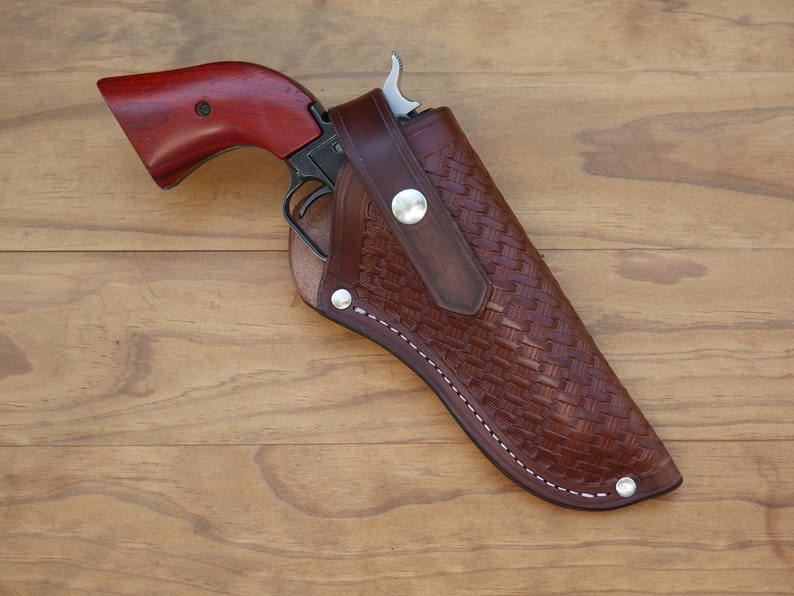 May include: A brown leather holster with a woven pattern holds a revolver with a dark metal frame and a reddish-brown wooden grip. The holster is secured with silver-colored rivets and white stitching.