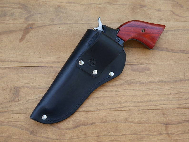 May include: A black leather holster with a silver-colored metal button closure, holding a revolver with a reddish-brown wooden grip. The revolver's trigger and hammer are visible. The holster is stitched with a curved design.