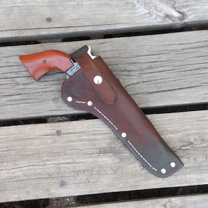 May include: A brown leather holster with a matching revolver. The holster is stitched with white thread and secured with silver rivets. The revolver has a wooden handle and a silver barrel. The items are displayed on a weathered wooden surface.