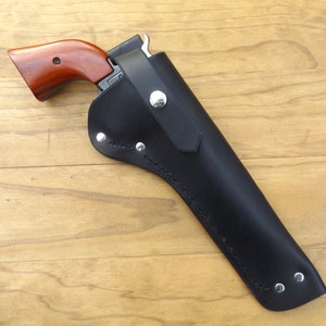 May include: A black leather holster with a silver button closure, holding a revolver with a brown wooden handle. The holster is riveted and stitched, designed to securely carry a firearm. The revolver is partially visible.