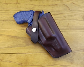 May include: A brown leather holster with a blue handgun. The holster has a strap with a silver snap closure. The handgun is partially visible, showing a blue grip. The items are on a wooden surface.