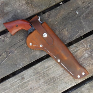 May include: A brown leather holster with a revolver. The holster has white stitching and silver rivets. The revolver has a wooden handle and a silver barrel. The items are on a wooden surface.