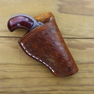 May include: A small handgun with a wood grip and a silver barrel is partially visible in a brown leather holster. The holster has a woven pattern and white stitching. The background is a wooden surface.