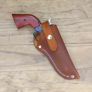 May include: A brown leather holster with a matching brown handle revolver. The holster has a stitched white border and a strap secured with a silver snap. The revolver has a wooden handle and a silver trigger.