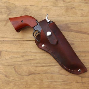 May include: A vintage revolver with a wooden handle is partially holstered in a brown leather case. The gun has a silver trigger and a dark metal frame. The holster is secured with a silver snap. The background is a light brown wooden surface.