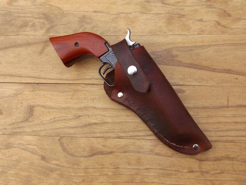 May include: A vintage revolver with a wooden handle is partially holstered in a brown leather case. The gun has a silver trigger and a dark metal frame. The holster is secured with a silver snap. The background is a light brown wooden surface.