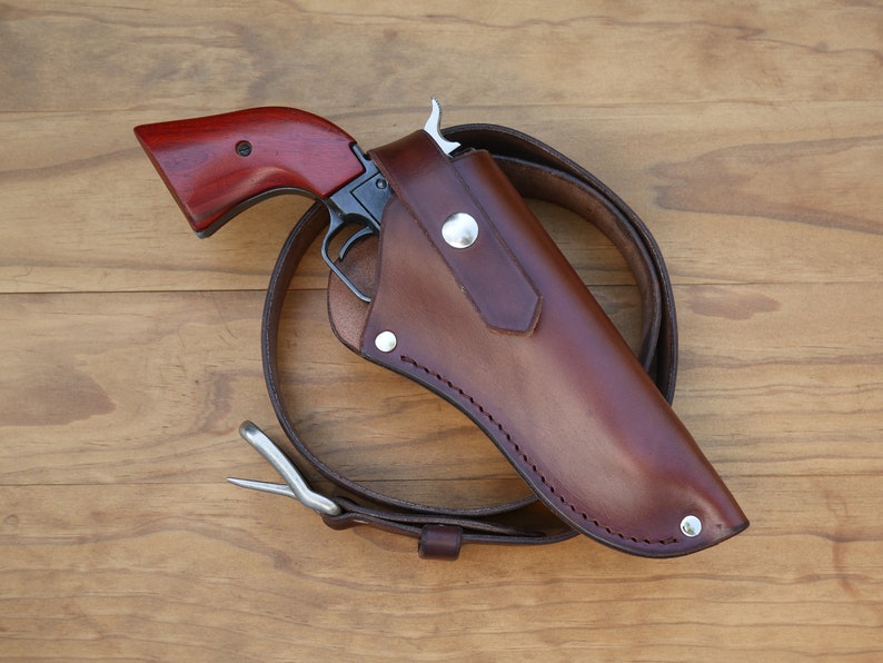 May include: A vintage-style handgun with a dark wooden grip is partially holstered in a brown leather holster. The holster is attached to a brown leather belt with a silver buckle. The gun and holster are set against a wooden background.