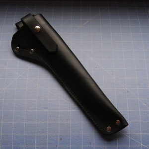 May include: A black leather sheath with a flap and snap closure. The sheath is designed to hold a tool, with rivets along the edges for reinforcement. The leather has a smooth, matte finish.
