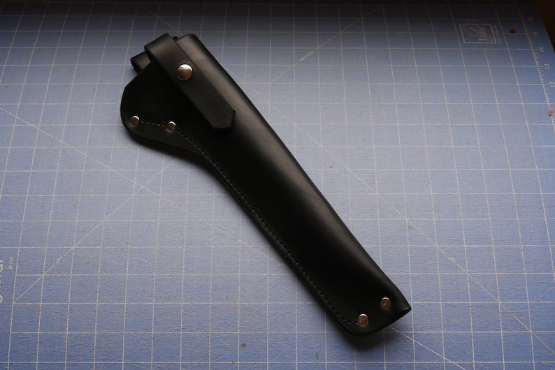 May include: A black leather sheath with a flap and snap closure. The sheath is designed to hold a tool, with rivets along the edges for reinforcement. The leather has a smooth, matte finish.