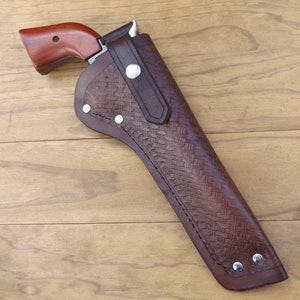 May include: A brown leather holster with a woven pattern, holding a revolver with a wooden grip. The holster has silver rivets and a belt loop. The revolver's grip is a reddish-brown color.