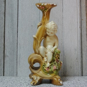 May include: A ceramic candle holder featuring a cherub figure sitting on a floral design. The holder is painted in shades of beige, brown, and green.