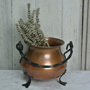 Peut inclure: Un chaudron en cuivre avec une texture martelée, des poignées en fer forgé noir et trois pieds incurvés. Le chaudron est rempli de lavande séchée. Le fond est un mur en bois de couleur claire.